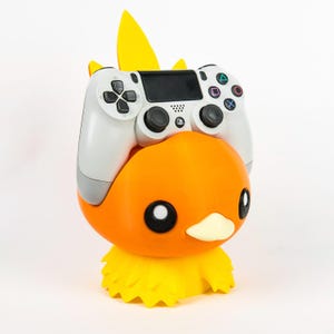 May include: An orange and yellow duck-shaped controller holder with a white PlayStation controller resting on top. The holder has black eyes and a yellow crest. The base is yellow and feathery.