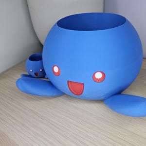 May include: A blue 3D printed planter shaped like a cartoon character with a smiling face and red eyes. The planter has a smaller planter attached to its side.