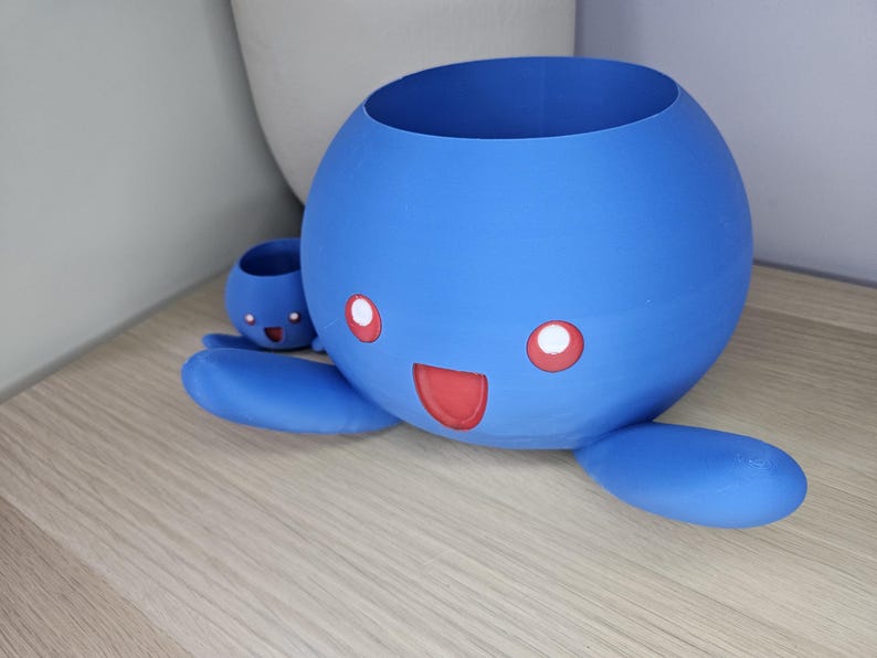 May include: A blue 3D printed planter shaped like a cartoon character with a smiling face and red eyes. The planter has a smaller planter attached to its side.