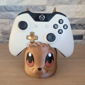 May include: A white Xbox controller with black accents and gold details rests on a brown, cartoonish animal-shaped stand. The controller has the standard button layout, including A, B, X, and Y buttons.