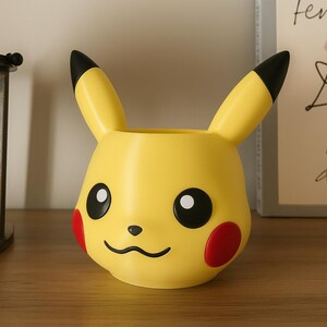 May include: A yellow Pikachu-shaped planter with black-tipped ears, black eyes, and red cheek circles. The planter has a wide opening for plants and is a decorative item for Pokemon fans.