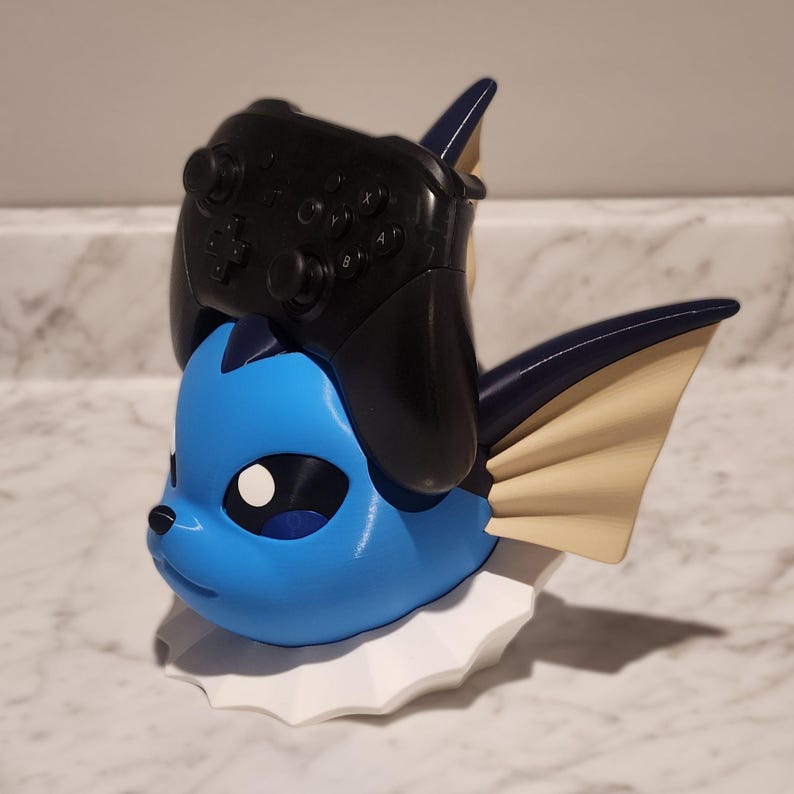 May include: A blue and white figurine of a cartoon character with a black controller resting on its head. The figurine has a white ruffled base and tan fins. The controller has black buttons and joysticks.