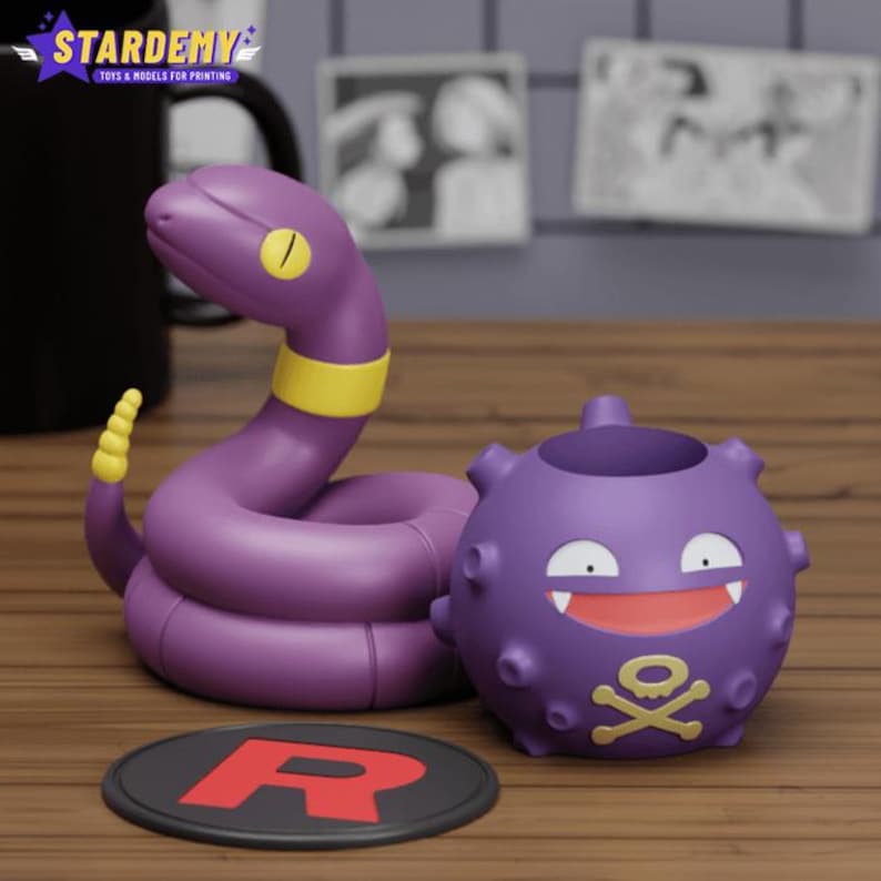 Puede incluir: Figuras de Pok&eacute;mon moradas y amarillas de una serpiente enroscada y una bola con pinchos con una calavera y huesos cruzados. Las figuras est&aacute;n sobre una base redonda negra con una R blanca.