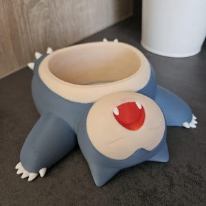 May include: A blue and white 3D printed planter shaped like a cartoon character with a wide open mouth and red lips. The planter is lying on its back.