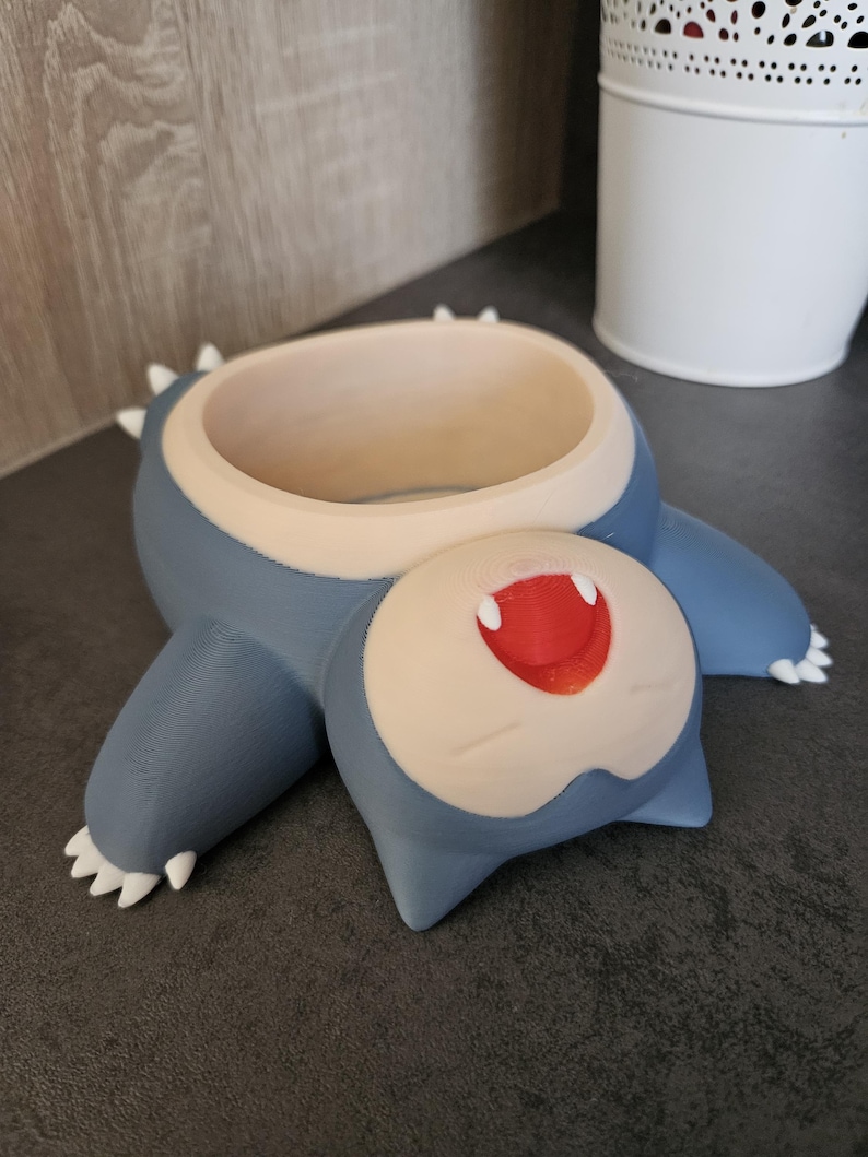 May include: A blue and white 3D printed planter shaped like a cartoon character with a wide open mouth and red lips. The planter is lying on its back.