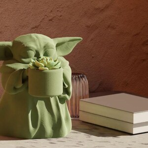 Puede incluir: Una figurita de cer&aacute;mica verde de un personaje de la franquicia Star Wars sosteniendo una peque&ntilde;a planta suculenta en maceta.