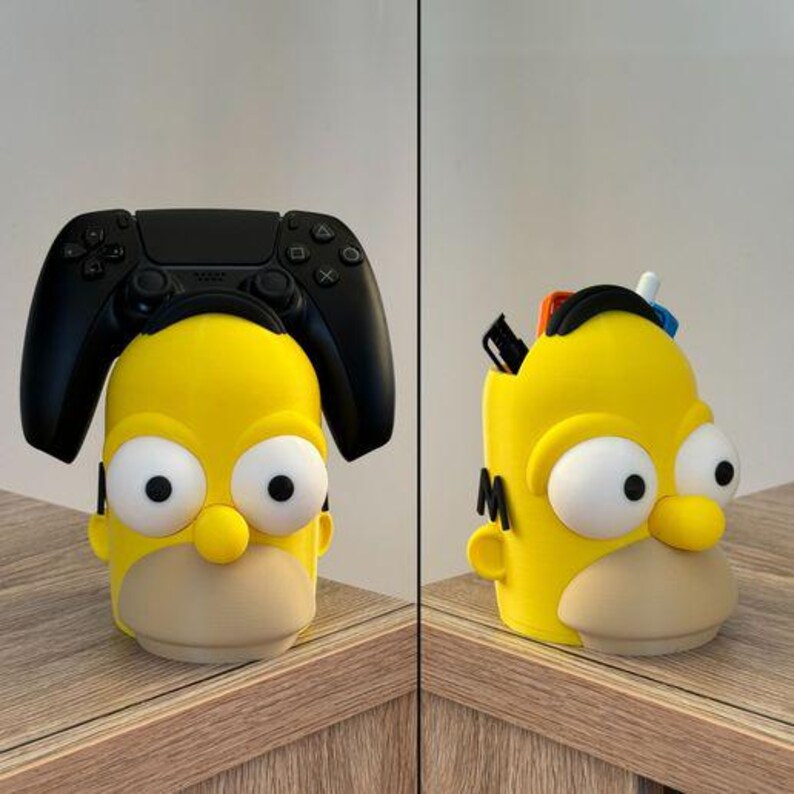 May include: A yellow and beige pen holder shaped like Homer Simpson's head, with large white eyes and a round nose. A black PlayStation controller rests on top of the pen holder. The second image shows the pen holder with pens and pencils inside.