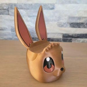 May include: A brown, 3D-printed Eevee-shaped planter with large ears and expressive eyes. The interior is hollow, suitable for small plants or decorative items. The ears have a pink interior.