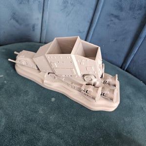 May include: A gray 3D printed model of a Star Wars AT-AT walker. The model is on a base and has a detailed design.