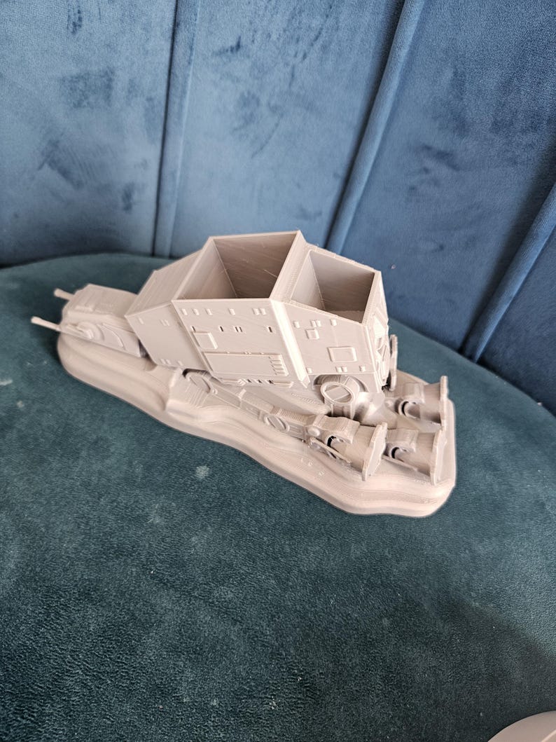 May include: A gray 3D printed model of a Star Wars AT-AT walker. The model is on a base and has a detailed design.