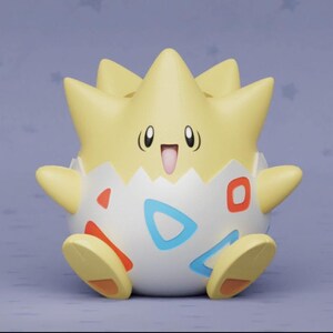 May include: A Togepi figurine, featuring a light yellow spiky head and arms, and a white egg-shaped body with red, blue, and orange accents. The figurine has a smiling face with black eyes and a pink mouth. The background is a light purple.