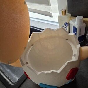 May include: A white, egg-shaped container with a hinged lid. The container has red and blue accents and is open, revealing a hollow interior. The lid is tan and has magnets. The container is on a dark surface.
