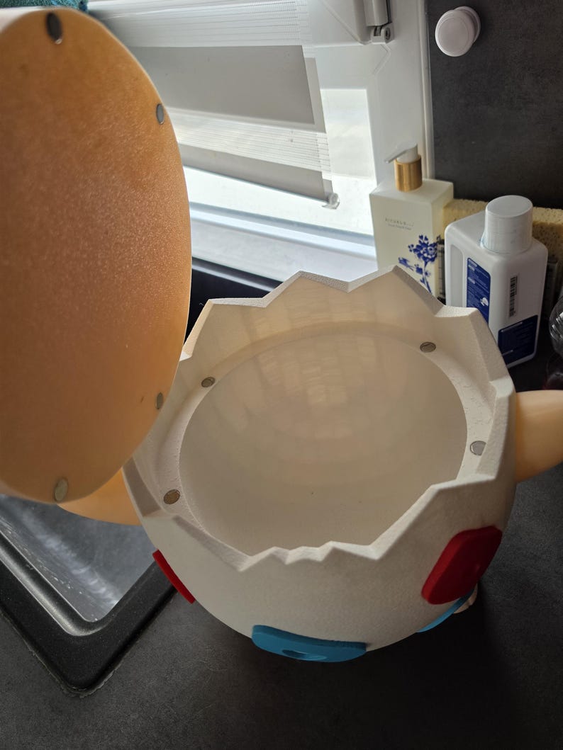 May include: A white, egg-shaped container with a hinged lid. The container has red and blue accents and is open, revealing a hollow interior. The lid is tan and has magnets. The container is on a dark surface.