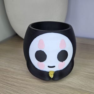 May include: Black and white ceramic planter shaped like a cartoon face with pink cheeks and black eyes. The planter has a small opening at the top for planting.