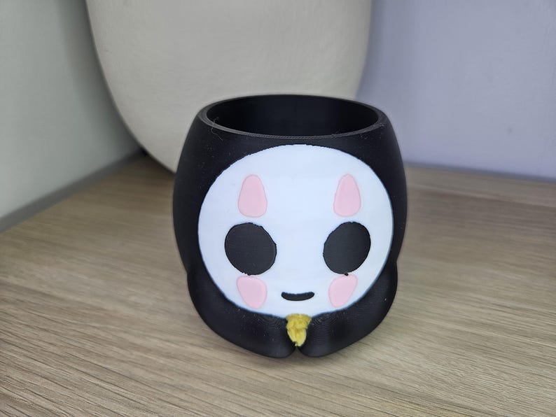 May include: Black and white ceramic planter shaped like a cartoon face with pink cheeks and black eyes. The planter has a small opening at the top for planting.