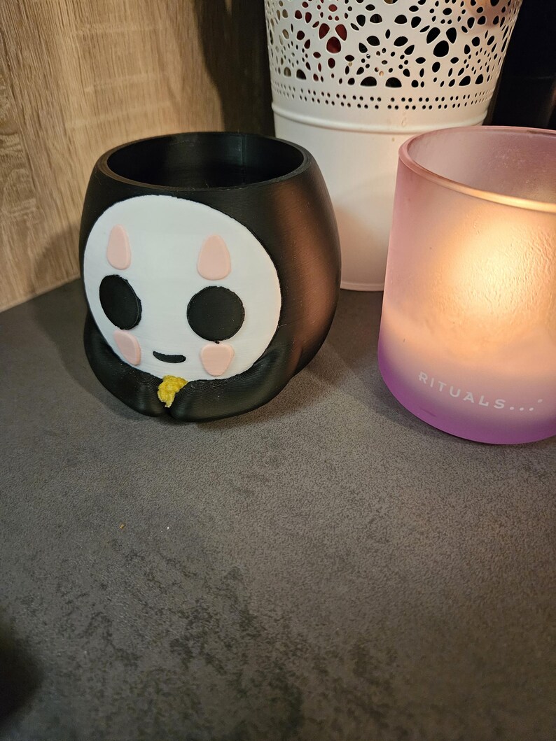 May include: Black and white ceramic planter shaped like a cartoon character with pink cheeks and black eyes. The planter is sitting on a brown surface.