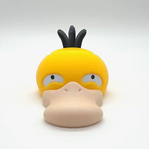 May include: A yellow Psyduck figure with a light beige beak and eyes. The figure has three black spikes on top of its head. The figure is a 3D-printed item.