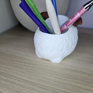 May include: White ceramic pen holder shaped like a fluffy animal with four legs. The holder has four pens inside, including a pink pen with a white design, a green pen with white stars, and two blue pens.