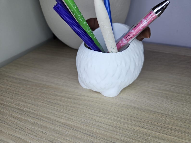 May include: White ceramic pen holder shaped like a fluffy animal with four legs. The holder has four pens inside, including a pink pen with a white design, a green pen with white stars, and two blue pens.