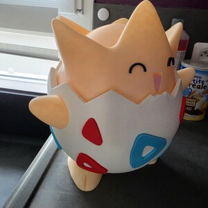 May include: A 3D-printed Togepi figure, a Pok&eacute;mon character, in a light orange colour. The figure is emerging from a white eggshell with red and blue accents. The figure has a smiling face with black eyes and a pink mouth.