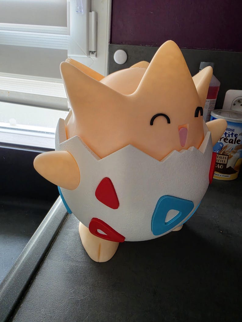 May include: A 3D-printed Togepi figure, a Pok&eacute;mon character, in a light orange colour. The figure is emerging from a white eggshell with red and blue accents. The figure has a smiling face with black eyes and a pink mouth.