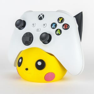 Pu&ograve; includere: Un controller Xbox bianco poggia su un supporto giallo a tema Pikachu. Il controller ha joystick neri e pulsanti colorati. Il supporto presenta un volto di Pikachu sorridente con cerchi sulle guance rosa e occhi neri.