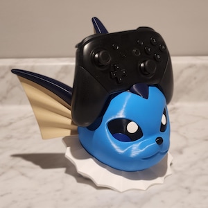May include: A blue and white 3D-printed Vaporeon figure with a black video game controller resting on top. The figure has a white frill and tan fins. The controller has black buttons and a directional pad.