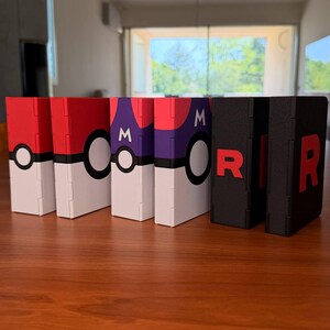 Puede incluir: Una colecci&oacute;n de cajas decorativas con varios dise&ntilde;os. Las cajas presentan esquemas de color rojo, blanco, morado y negro. Algunas cajas tienen un dise&ntilde;o de Pok&eacute; Ball, mientras que otras muestran la letra "R". Las cajas est&aacute;n dispuestas sobre una superficie de madera.