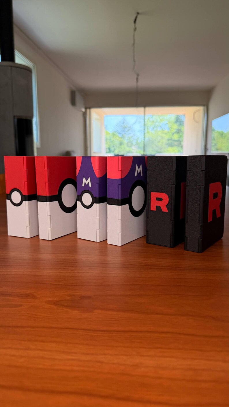 Puede incluir: Una colecci&oacute;n de cajas decorativas con varios dise&ntilde;os. Las cajas presentan esquemas de color rojo, blanco, morado y negro. Algunas cajas tienen un dise&ntilde;o de Pok&eacute; Ball, mientras que otras muestran la letra "R". Las cajas est&aacute;n dispuestas sobre una superficie de madera.