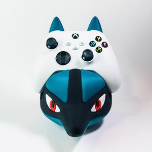 Puede incluir: Un mando Xbox blanco descansa sobre una base azul y negra con temática de Lucario. El mando tiene joysticks negros, un panel direccional y botones de acción de colores. La base presenta ojos rojos y un diseño estilizado.