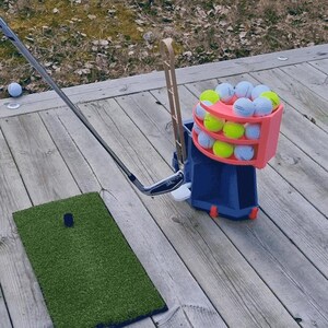 May include: A golf practice setup featuring a blue and pink ball dispenser filled with golf balls, a golf club, a green turf mat, and a wooden club holder. The setup is on a wooden deck, with a golf ball resting on the deck.
