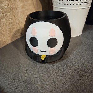 May include: Black and white ceramic planter shaped like a cartoon face with pink cheeks and black eyes. The planter has a yellow stem.