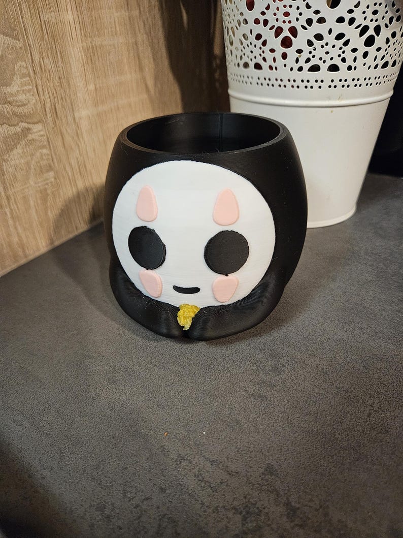 May include: Black and white ceramic planter shaped like a cartoon face with pink cheeks and black eyes. The planter has a yellow stem.
