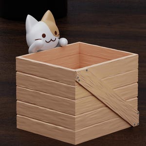 May include: A wooden crate with a cat figurine peeking out of the top. The crate is made of light brown wood with a diagonal brace across the front. The cat is white with brown markings and has a cute expression.