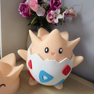 May include: A light tan Togepi figure, a Pok&eacute;mon character, is shown in the image. The figure has a white eggshell, decorated with red and blue shapes. Another Togepi figure is partially visible to the left. A bouquet of pink and purple flowers is in the background.