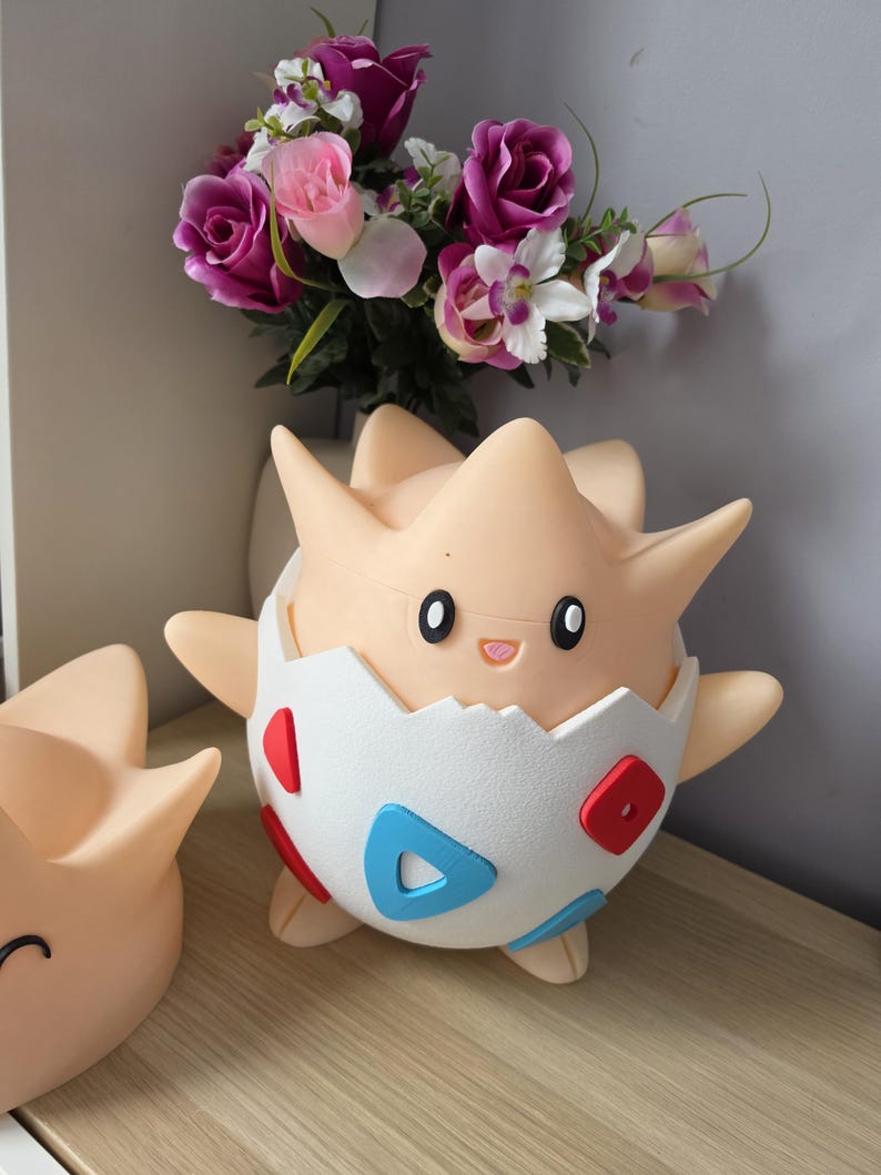 May include: A light tan Togepi figure, a Pok&eacute;mon character, is shown in the image. The figure has a white eggshell, decorated with red and blue shapes. Another Togepi figure is partially visible to the left. A bouquet of pink and purple flowers is in the background.