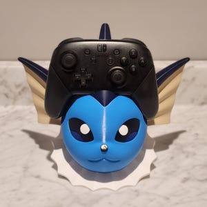 May include: A blue and white decorative stand shaped like a cartoon character, holding a black Nintendo Switch controller. The stand has a white base and blue accents, with a detailed face and ears.