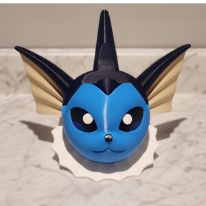 May include: A 3D-printed Vaporeon head, with a light blue face, black ears, and a dorsal fin. The design includes tan fins and white accents, mounted on a white, scalloped base. The sculpture is a fan-made item.