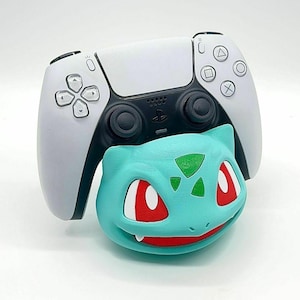 May include: A white and black PlayStation 5 controller rests above a teal Bulbasaur-shaped controller holder. The Bulbasaur has red eyes and a green patch on its forehead. The controller has the standard button layout.