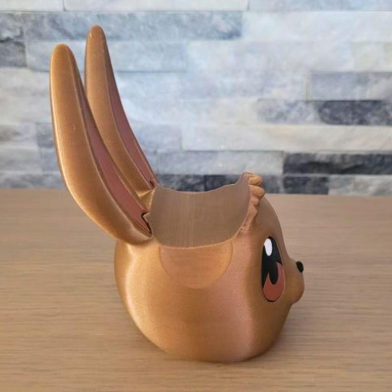 May include: A 3D-printed planter in the shape of a cartoon character's head, featuring large ears and a round body. The planter is primarily a light brown color with pink accents inside the ears and around the eyes. The eyes are black and white.