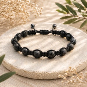 Könnte beinhalten: Ein schwarzes Armband mit einer Reihe von porösen, runden Lavasteinperlen, die durch eine geflochtene schwarze Kordel verbunden sind. Das Design verfügt über einen verstellbaren Schiebeknotenverschluss mit zwei kleinen schwarzen Perlen an den Enden.