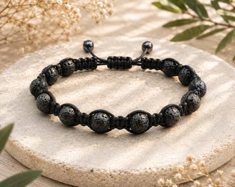 May include: A black bracelet featuring a series of porous, round lava stone beads connected by a woven black cord. The design includes an adjustable sliding knot closure with two small black beads at the ends of the cord strings for a secure fit.