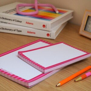 Op de afbeelding: Twee witte notitieblokken met roze gestreepte randen en een roze rand, gestapeld op een houten oppervlak. Twee oranje potloden en een roze pen liggen naast de notitieblokken. Boeken en een roze bureaulamp op de achtergrond.