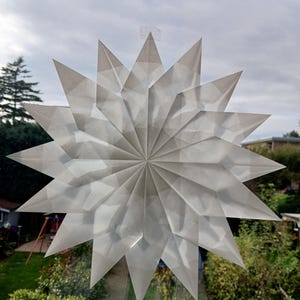 May include: A large, white paper star decoration is attached to a window. The star has a complex, folded paper design with many pointed edges. The background shows a cloudy sky and green trees and bushes.