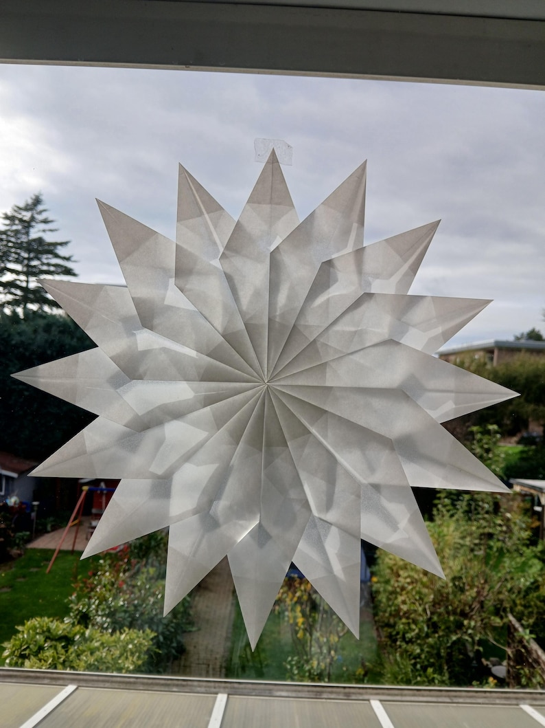 May include: A large, white paper star decoration is attached to a window. The star has a complex, folded paper design with many pointed edges. The background shows a cloudy sky and green trees and bushes.