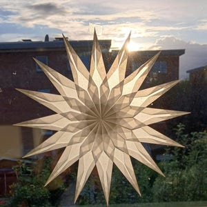 May include: A large, paper star decoration with a geometric design is affixed to a window. The star is a light beige colour and has many pointed edges. The sun shines through the star, creating a bright, glowing effect. The background includes a house and trees.
