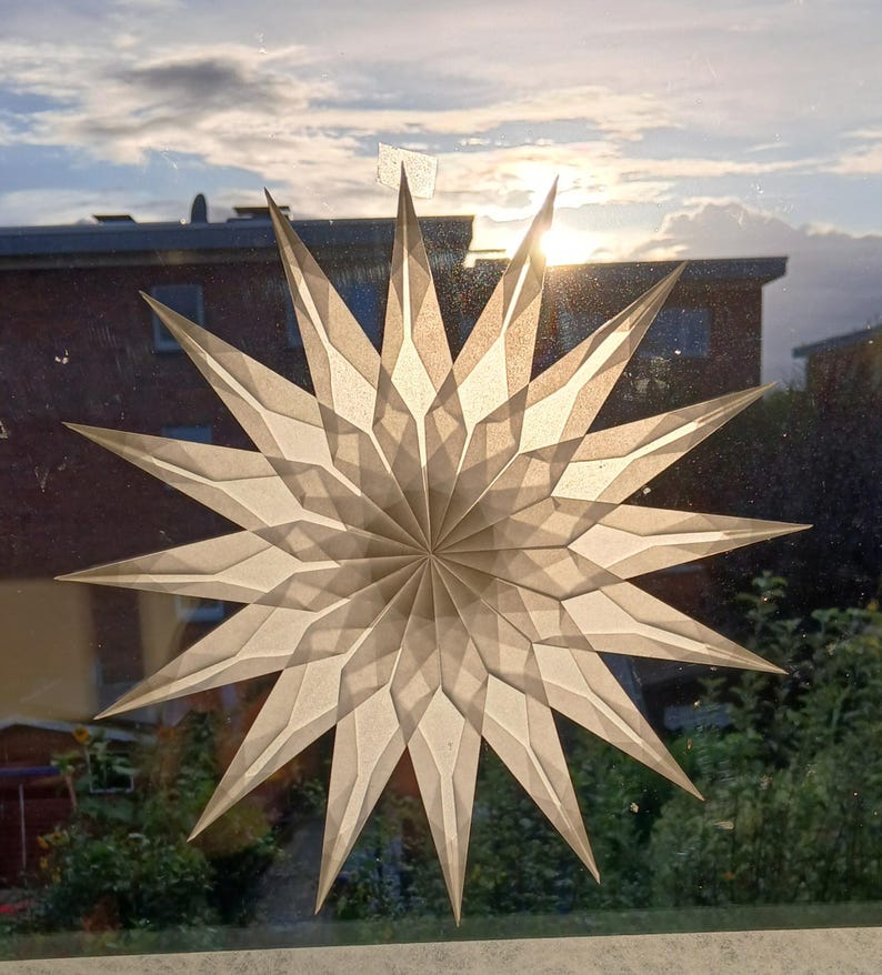 May include: A large, paper star decoration with a geometric design is affixed to a window. The star is a light beige colour and has many pointed edges. The sun shines through the star, creating a bright, glowing effect. The background includes a house and trees.