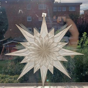 May include: A large, white paper star decoration with multiple points, attached to a window. The star is made of folded paper, creating a layered effect. The background shows buildings and greenery.