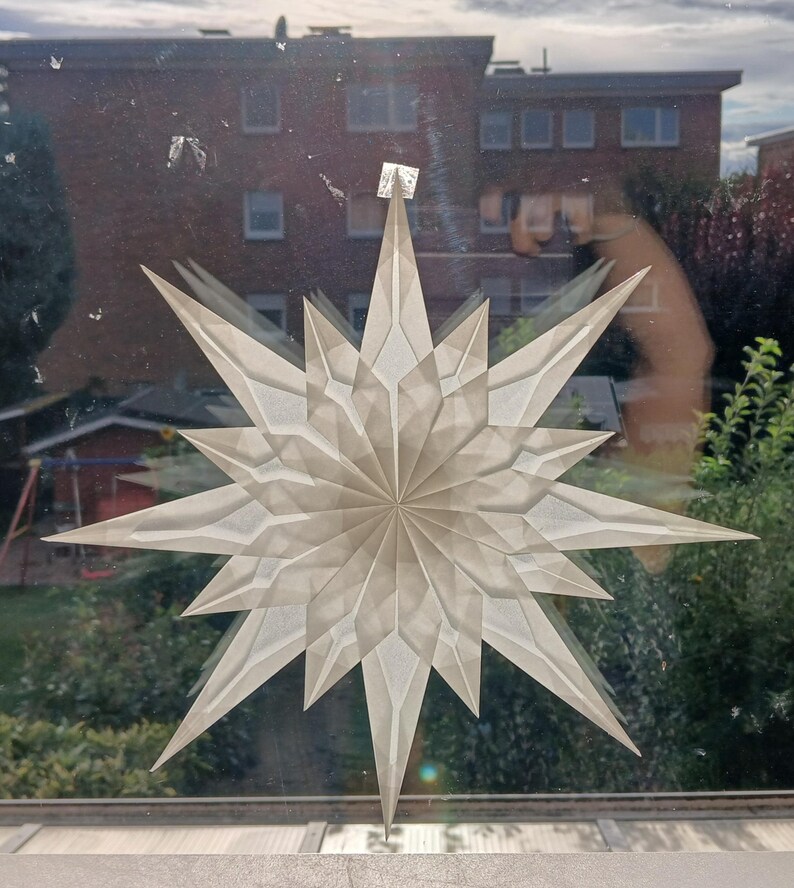 May include: A large, white paper star decoration with multiple points, attached to a window. The star is made of folded paper, creating a layered effect. The background shows buildings and greenery.
