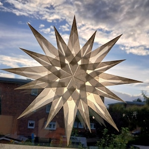 May include: A decorative star, made from translucent paper, attached to a window. The star has multiple points and a geometric design. The background shows a cloudy sky and a building. The star's design allows light to pass through.